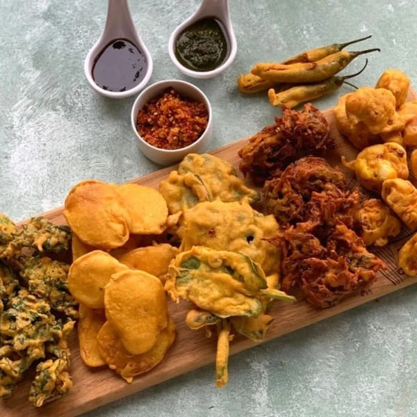 A wooden board filled with various Indian pakora, including chili, onion, and spinach, with dipping sauces.